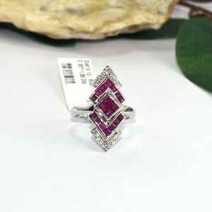 Puede incluir: Un anillo de plata con un diseño geométrico alargado, engastado con múltiples rubíes de color rosa intenso de corte cuadrado y pequeños diamantes blancos redondos. El anillo incluye una etiqueta blanca con texto impreso.