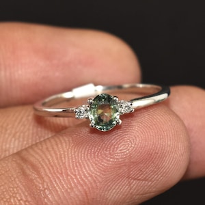 Op de afbeelding: Een zilveren ring met een ovale groene edelsteen in het midden, geflankeerd door twee kleinere witte edelstenen. De ring wordt op een vinger gepresenteerd tegen een donkere achtergrond, wat het delicate ontwerp en de glans van het sieraad benadrukt.