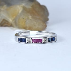 Puede incluir: Un anillo de banda de plata con una fila de zafiros azules, rubíes rojos y un solo diamante en el centro.