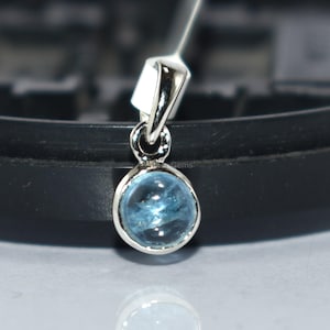 May include: A small, round, light blue gemstone pendant with a silver-colored setting. The pendant is suspended from a thin, silver-colored chain. The gemstone is approximately 0.3 inches in diameter. The pendant is displayed against a blurred black background.