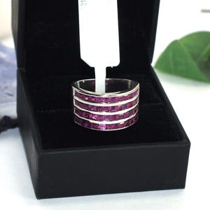 May include: A silver-toned ring featuring four horizontal rows of square-cut, deep magenta-colored stones set in a channel setting. The ring is displayed inside a black jewelry box with a white rectangular price tag attached to the band.
