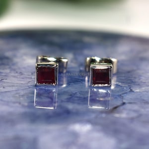 Puede incluir: Un par de pendientes de botón cuadrados con piedras preciosas de color rojo oscuro engastadas en plata. Los pendientes se muestran sobre una superficie azul texturizada. Pequeños y elegantes, son adecuados para el uso diario o para ocasiones especiales.