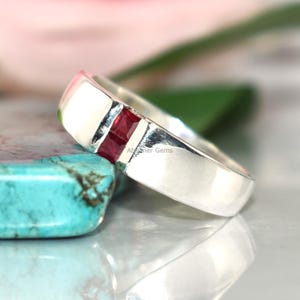 May include: A silver ring with a rectangular, deep red gemstone inlay. The ring is set against a blurred background with a turquoise stone and pink flower, highlighting the jewelry's design and color.