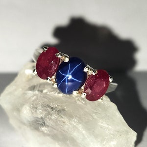 May include: A silver ring featuring three gemstones. The center stone is a blue star sapphire, flanked by two oval-cut ruby gemstones. The ring is set on a white, crystalline surface, with a shadow in the background.