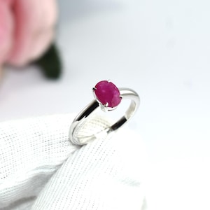 Puede incluir: Un anillo de tono plateado con una gema única de corte ovalado en color rojo rosado intenso, montada en cuatro garras. La banda es lisa y pulida, mostrando un acabado metálico limpio que complementa el color vibrante de la piedra central.
