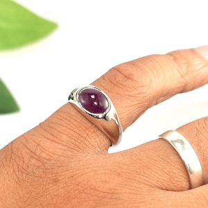 May include: A silver ring featuring a smooth, oval-shaped deep purple cabochon gemstone set in a polished metal band, worn alongside a plain, solid silver band on the adjacent finger, showcasing a clean, metallic finish and a rounded stone.