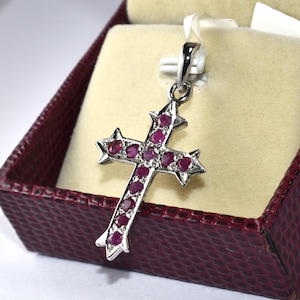 Könnte beinhalten: Ein silberner Kreuzanhänger mit Sternmuster an den Enden jedes Arms. Das Kreuz ist mit zahlreichen kleinen, runden, tiefroten Edelsteinen besetzt. Der Anhänger wird in einer bordeauxfarbenen Schmuckschatulle mit cremefarbenem Innenraum präsentiert.