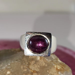 May include: A silver ring with a square setting holding a large, oval-shaped, deep red gemstone. The ring is set on a light-colored, textured surface. The gemstone has a polished, reflective surface.