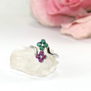 Può includere: Un anello color argento caratterizzato da due grappoli di piccole pietre preziose a forma di fiore. Il grappolo superiore è composto da quattro pietre rotonde verdi, mentre quello inferiore da quattro pietre rotonde magenta su una fascia sottile.