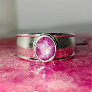 Op de afbeelding: Een zilveren ring met een roze edelsteen in een bezel-setting. De ring heeft een brede band met een getextureerd oppervlak.
