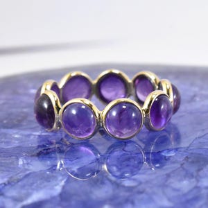 Könnte beinhalten: Ein Goldring mit einer durchgehenden Reihe runder, im Cabochon-Schliff gehaltener violetter Amethyst-Edelsteine. Die Steine sind in einzelnen Goldfassungen gefasst, die zu einem kreisförmigen Band verbunden sind und eine polierte Oberfläche aufweisen.