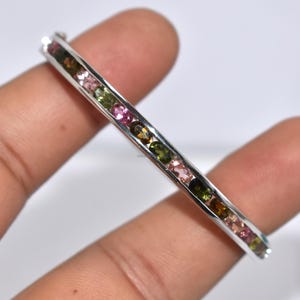 May include: A silver bracelet featuring a line of multi-coloured gemstones. The gems are pink, green, and brown, arranged in a row. The bracelet is held in a hand.