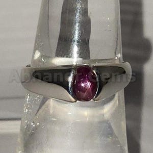 Puede incluir: Un anillo de plata con una gran gema ovalada de color rosa intenso. El anillo tiene un diseño simple y moderno con un acabado pulido. La gema es el punto focal, engastada en un bisel. El anillo se exhibe sobre un soporte transparente.