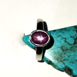 May include: A silver ring with a large, oval, star ruby gemstone. The ruby is a deep pink color with a six-ray star pattern. The ring is set on a turquoise-colored surface.