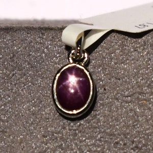 May include: An oval pendant featuring a star ruby gemstone. The ruby is a deep reddish-purple hue, set in a gold-coloured metal frame. The pendant hangs from a small loop, and a white tag is attached.