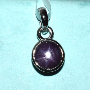 Peut inclure: Pendentif avec une pierre précieuse ronde, violet foncé, présentant un motif en étoile. La pierre est sertie dans une lunette en métal argenté et fixée à une bélière argentée.