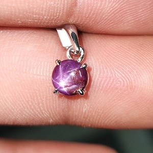 May include: A small, round, purple pendant with a star-like effect. The gemstone is set in a silver-colored metal setting with a small loop for a chain. The pendant is held between fingers, showcasing its size and color.