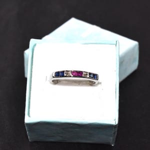 K&ouml;nnte beinhalten: Ein silberner Bandring mit blauen und rosafarbenen Edelsteinen. Der Ring hat auf jeder Seite eines gr&ouml;&szlig;eren rosafarbenen Edelsteins eine Reihe kleiner blauer Edelsteine.