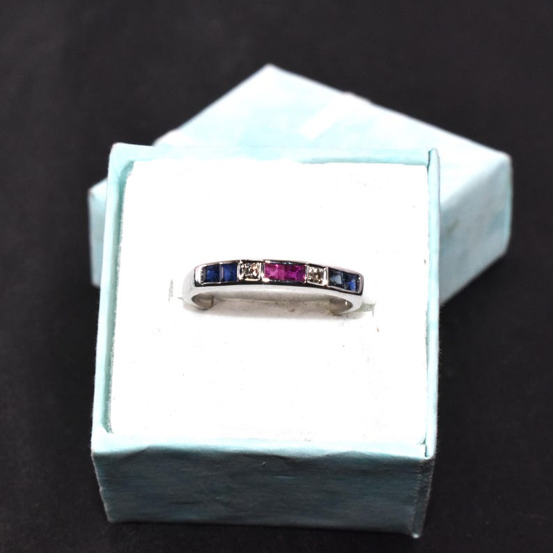 K&ouml;nnte beinhalten: Ein silberner Bandring mit blauen und rosafarbenen Edelsteinen. Der Ring hat auf jeder Seite eines gr&ouml;&szlig;eren rosafarbenen Edelsteins eine Reihe kleiner blauer Edelsteine.