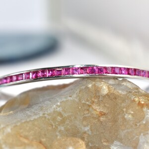 May include: A silver bracelet featuring a line of square-cut, bright pink gemstones. The bracelet is displayed on a light-coloured stone, with a blurred background.