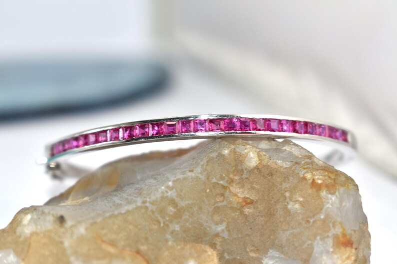 May include: A silver bracelet featuring a line of square-cut, bright pink gemstones. The bracelet is displayed on a light-coloured stone, with a blurred background.