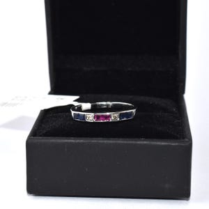 K&ouml;nnte beinhalten: Ein Wei&szlig;gold-Bandring mit einer Reihe blauer Saphire und einem einzelnen Rubin in der Mitte. Der Ring befindet sich in einer schwarzen Ringbox.