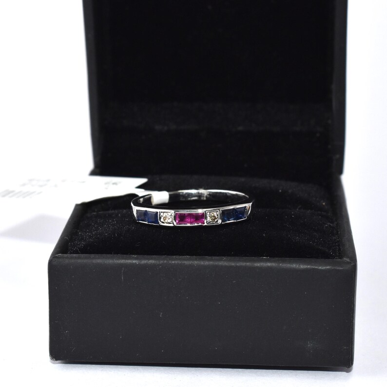 K&ouml;nnte beinhalten: Ein Wei&szlig;gold-Bandring mit einer Reihe blauer Saphire und einem einzelnen Rubin in der Mitte. Der Ring befindet sich in einer schwarzen Ringbox.