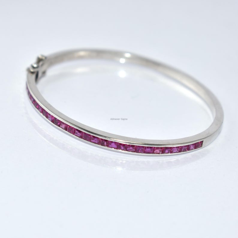 May include: A silver bangle bracelet featuring a line of square-cut, bright pink gemstones. The bracelet has a simple, elegant design with a clasp closure. The gemstones are set closely together, creating a continuous line of colour.