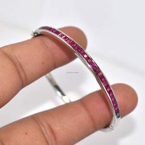 May include: A silver bangle bracelet featuring a line of square-cut, bright pink gemstones. The bracelet is held by a hand against a white backdrop, highlighting the polished metal and the colour of the gemstones. The bracelet is a piece of jewellery.