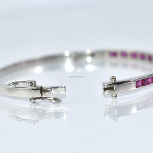 May include: Open silver bracelet with a row of square-cut pink gemstones. The bracelet is unclasped, showing the clasp mechanism. The gemstones are set in a channel setting. The bracelet is on a white background.