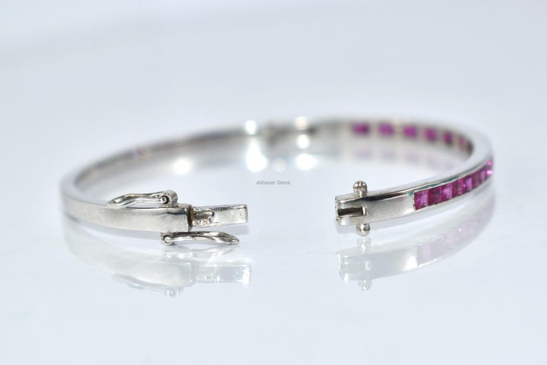 May include: Open silver bracelet with a row of square-cut pink gemstones. The bracelet is unclasped, showing the clasp mechanism. The gemstones are set in a channel setting. The bracelet is on a white background.