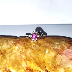 May include: A silver ring with a pink star sapphire gemstone. The ring has a delicate, curved design.