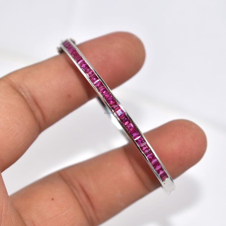 May include: A silver bangle bracelet featuring a line of square-cut, bright pink gemstones. The gemstones are set closely together, forming a continuous band of colour. The bracelet is held by a hand against a white background.
