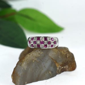 Könnte beinhalten: Ein silberfarbener Ring mit einem Schachbrettmuster aus abwechselnd quadratisch geschliffenen roten Rubinen und klaren weißen Diamanten. Die Ringschiene hat eine glatte, polierte Oberfläche und ist auf der Vorderseite mit mehreren Steinreihen besetzt.