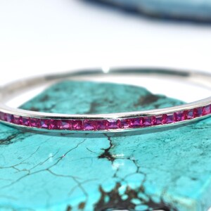 May include: A silver bangle bracelet featuring a line of square, vivid pink gemstones. The bracelet is displayed on a turquoise stone against a white backdrop. This elegant jewellery piece is suitable for special events.