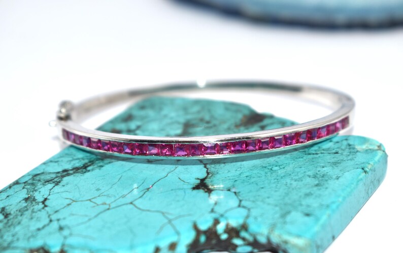 May include: A silver bangle bracelet featuring a line of square, vivid pink gemstones. The bracelet is displayed on a turquoise stone against a white backdrop. This elegant jewellery piece is suitable for special events.