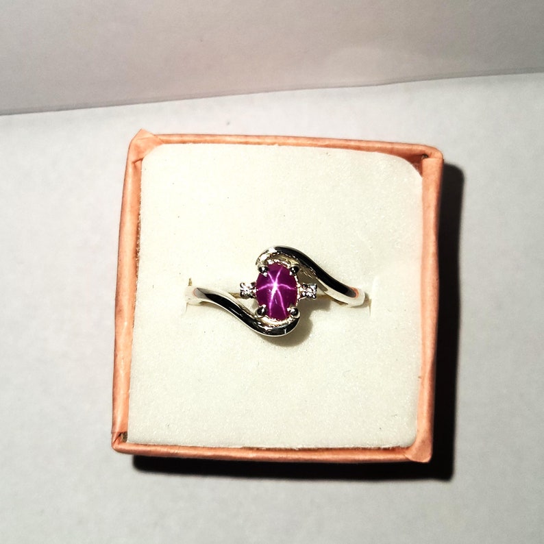 May include: A silver ring with a pink star ruby gemstone and two smaller diamonds. The ring is in a white box with a pink lid.