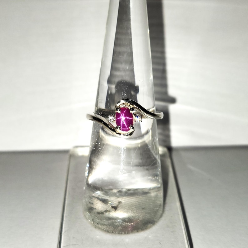 May include: A silver ring with a pink star ruby gemstone and two smaller diamonds. The ring is on a clear ring display stand.