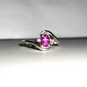 May include: A silver ring with a pink star ruby gemstone and two smaller diamonds. The ring has a curved band.