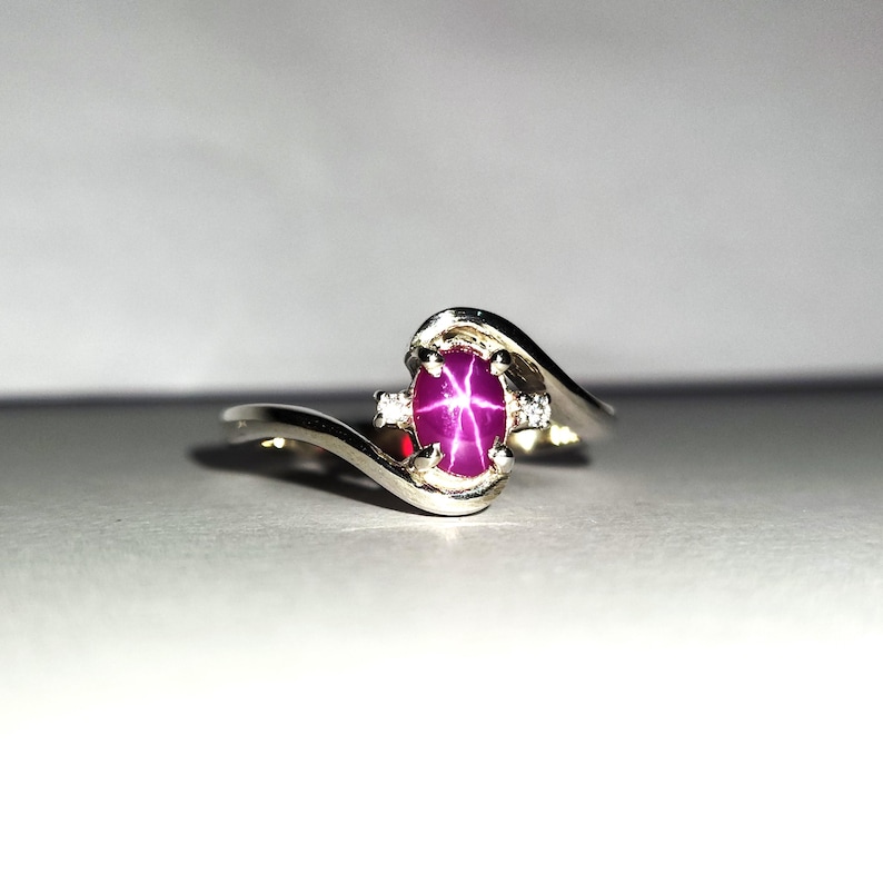 May include: A silver ring with a pink star ruby gemstone and two smaller diamonds. The ring has a curved band.