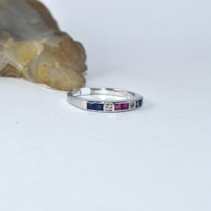 K&ouml;nnte beinhalten: Ein Silberring mit einer Reihe kleiner quadratischer Edelsteine. Die Edelsteine sind blau, klar und rot.