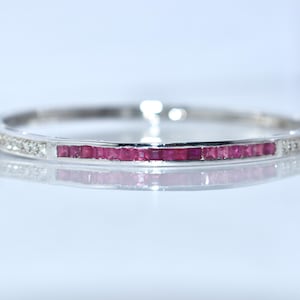 Puede incluir: Una pulsera de plata con una fila central de piedras preciosas rojas cuadradas. La pulsera también está adornada con pequeñas gemas claras a lo largo de los bordes, creando un efecto brillante. La joya se exhibe sobre una superficie reflectante.