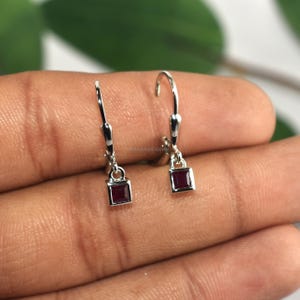 May include: A pair of silver leverback earrings. Each earring features a small, square, deep red gemstone suspended from a leverback closure. The earrings are displayed against a neutral background.