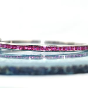May include: A silver bangle bracelet featuring a line of square-cut, bright pink gemstones. The bracelet is displayed on a light blue surface, reflecting the design. The gemstones are a vivid, striking colour.