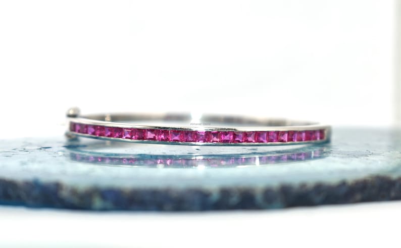 May include: A silver bangle bracelet featuring a line of square-cut, bright pink gemstones. The bracelet is displayed on a light blue surface, reflecting the design. The gemstones are a vivid, striking colour.