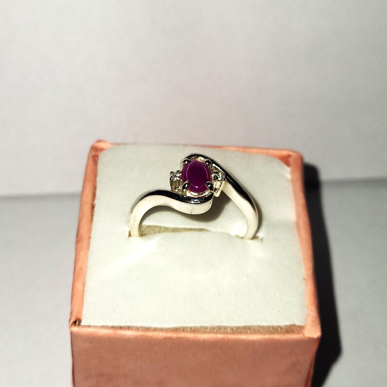 May include: A silver ring with a purple oval gemstone and two smaller clear gemstones. The ring is set on a white background.