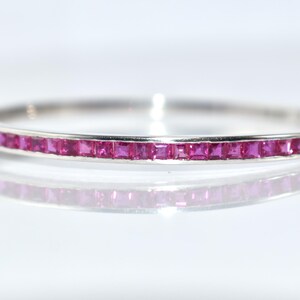 May include: A silver bangle bracelet featuring a line of square-cut, bright pink gemstones. The bracelet is set against a white backdrop, with its reflection below. The clasp is visible on the left. Jewellery.