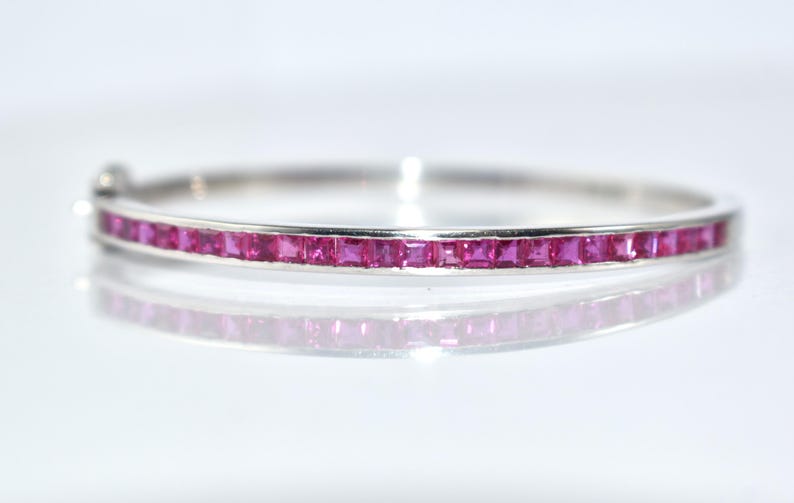 May include: A silver bangle bracelet featuring a line of square-cut, bright pink gemstones. The bracelet is set against a white backdrop, with its reflection below. The clasp is visible on the left. Jewellery.