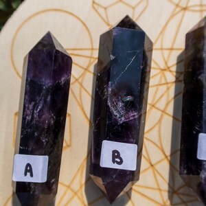 May include: Three dark purple crystal wands with pointed ends, labeled A, B, and C, are arranged on a wooden surface with a geometric pattern.