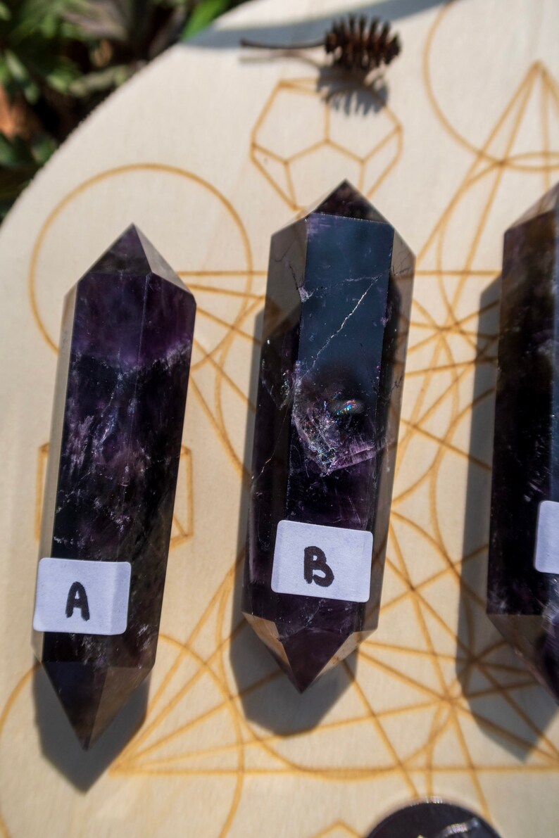 May include: Three dark purple crystal wands with pointed ends, labeled A, B, and C, are arranged on a wooden surface with a geometric pattern.